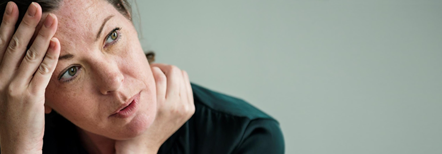 Photograph of a woman, listening and looking to one side. Her hand is touching her forehead. She looks tired, possibly unwell. She has light skin, dark haair and apprears to be in her 20's. She is wearing a dark green sweater.