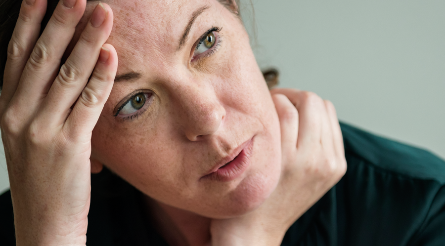 Woman listening hard. Head in hand. She looks stressed. She has dark hair, hazel eyes and her hair is tied back.