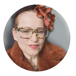 Image of a woman, head and shoulders. She is looking at us, and wearing a jacket and hat in autumn colours. She is also wearing bold glasses in brown. She has blue eyes and reddish hair, tied back.