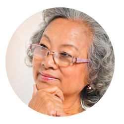 Image of an older Asian woman with wavy grey hair. She is wearing glasses and smiling thoughtfully. She is facing the left and looking over that way. Her hand is next to her face. She looks kind.